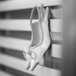 Par de zapatos de novia de tacón con detalles brillantes colgados sobre una superficie de madera, en una fotografía en blanco y negro. Imagen elegante y artística, ideal para una invitación de boda con estilo sofisticado y atemporal.