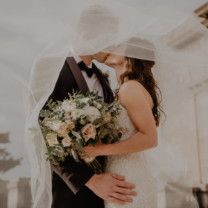 Pareja de recién casados compartiendo un beso cubiertos por el velo de la novia, mientras ella sostiene un ramo de flores en tonos crema y verdes. Escena romántica y delicada, perfecta para una invitación de boda elegante y emocional.