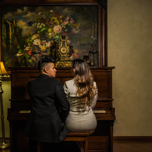 Pareja sentada de espaldas frente a un piano antiguo, en un salón decorado con arte clásico y una pintura floral. Él viste traje oscuro y ella un vestido blanco ajustado. Imagen perfecta para una invitación de boda con estilo minimalista, íntimo y sofisticado.