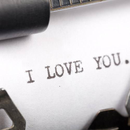 Primer plano de una hoja de papel en máquina de escribir con el texto “I LOVE YOU.” escrito en letras mayúsculas, enmarcado dentro de un círculo sobre fondo beige claro. Imagen con estilo vintage y romántico.