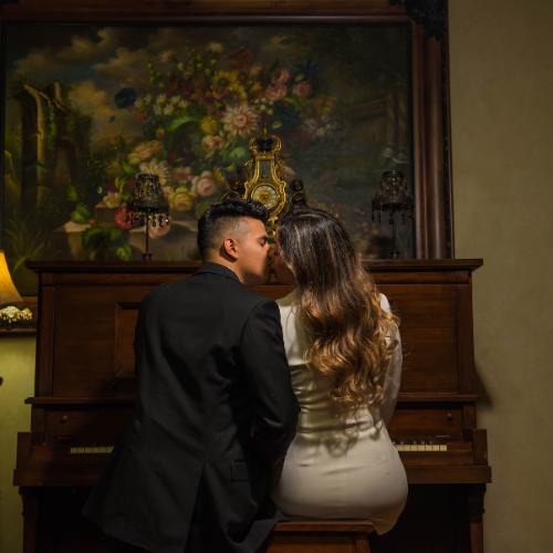 Pareja sentada frente a un piano, compartiendo un beso en un ambiente clásico con iluminación cálida y una pintura floral de fondo. Escena romántica y sobria, ideal para una invitación de boda minimalista con un toque artístico y elegante.