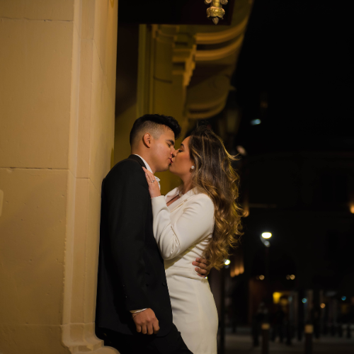 Pareja vestida con estilo formal besándose bajo la luz cálida de una farola en una calle nocturna. Ella luce un vestido blanco ajustado y él un traje oscuro. Imagen perfecta para una invitación de boda minimalista que combine elegancia, pasión y un ambiente urbano sofisticado.