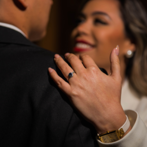 Primer plano de una mujer abrazando a su pareja mientras muestra un anillo de compromiso con piedra azul en su mano. Ella sonríe con emoción, representando un momento íntimo y especial, ideal para una invitación de boda o anuncio de compromiso sofisticado.
