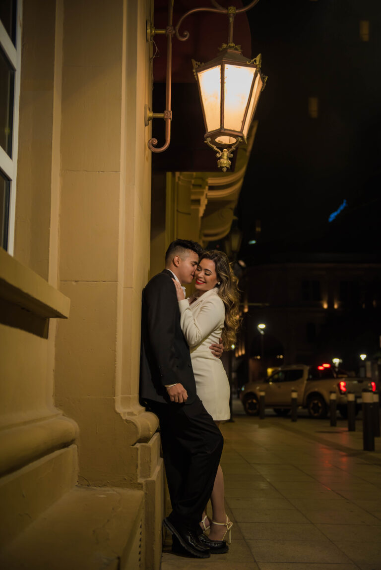 Pareja vestida de forma elegante abrazándose bajo una farola antigua en una calle iluminada por la noche. Escena íntima y sofisticada, perfecta para una invitación de boda con estilo urbano, moderno o nocturno.