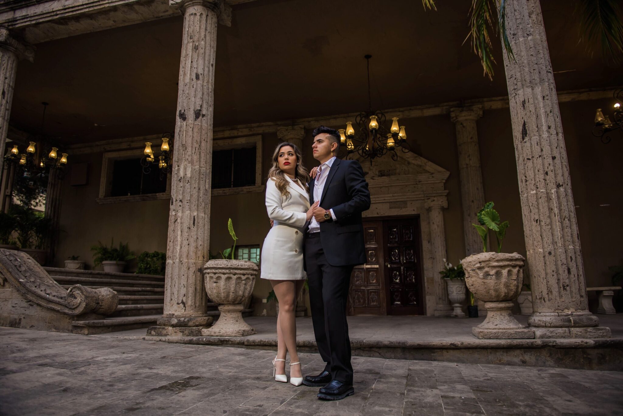 Pareja posando de manera formal frente a una edificación con columnas talladas y lámparas colgantes. Ella lleva vestido blanco tipo blazer y él traje oscuro. Imagen ideal para una invitación de boda minimalista con un entorno arquitectónico imponente y sofisticado.